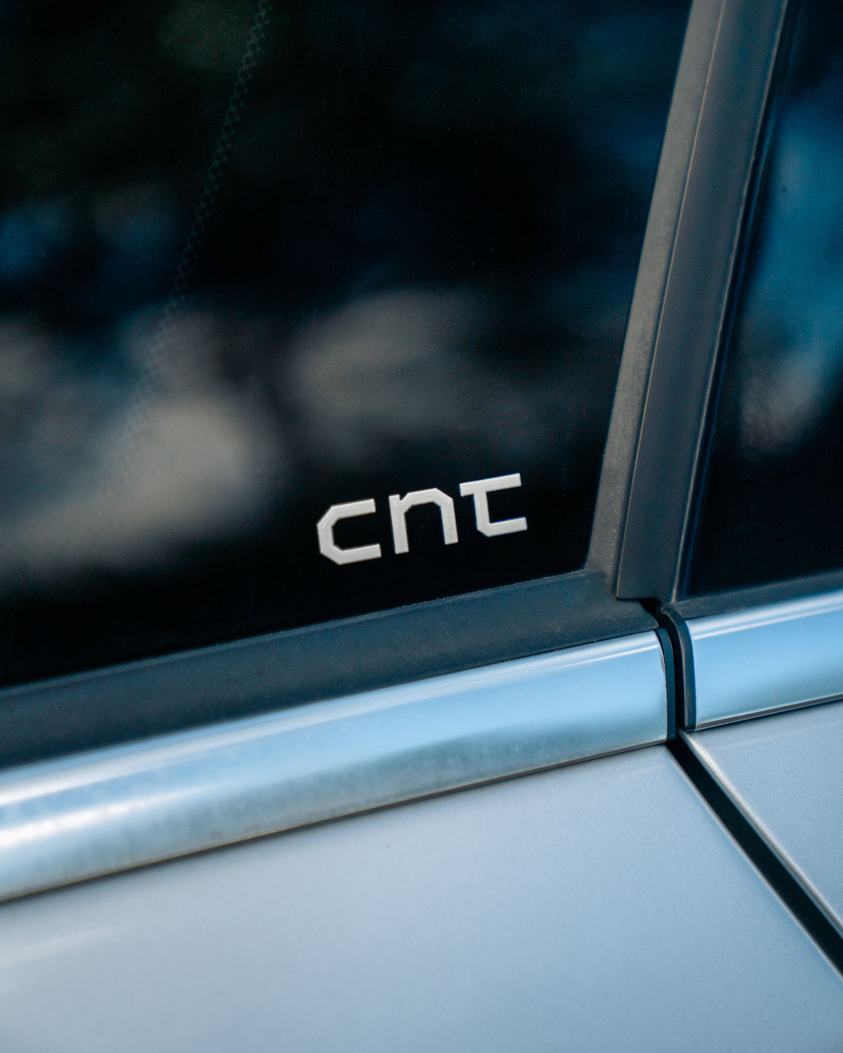 A close-up shot of a silver metallic 'cnt' emblem decal applied to the dark tinted window of a silver vehicle.