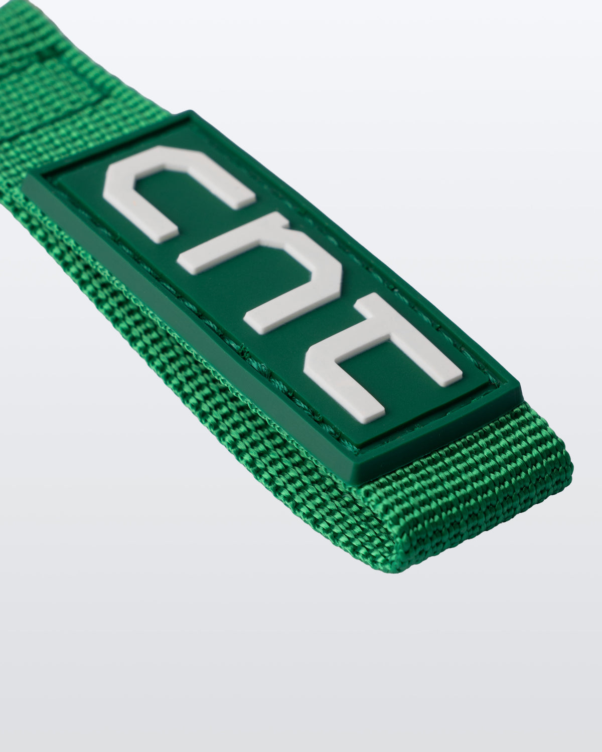A green woven strap with a rectangular green patch featuring large white stylized letters, photographed on a light gray background.