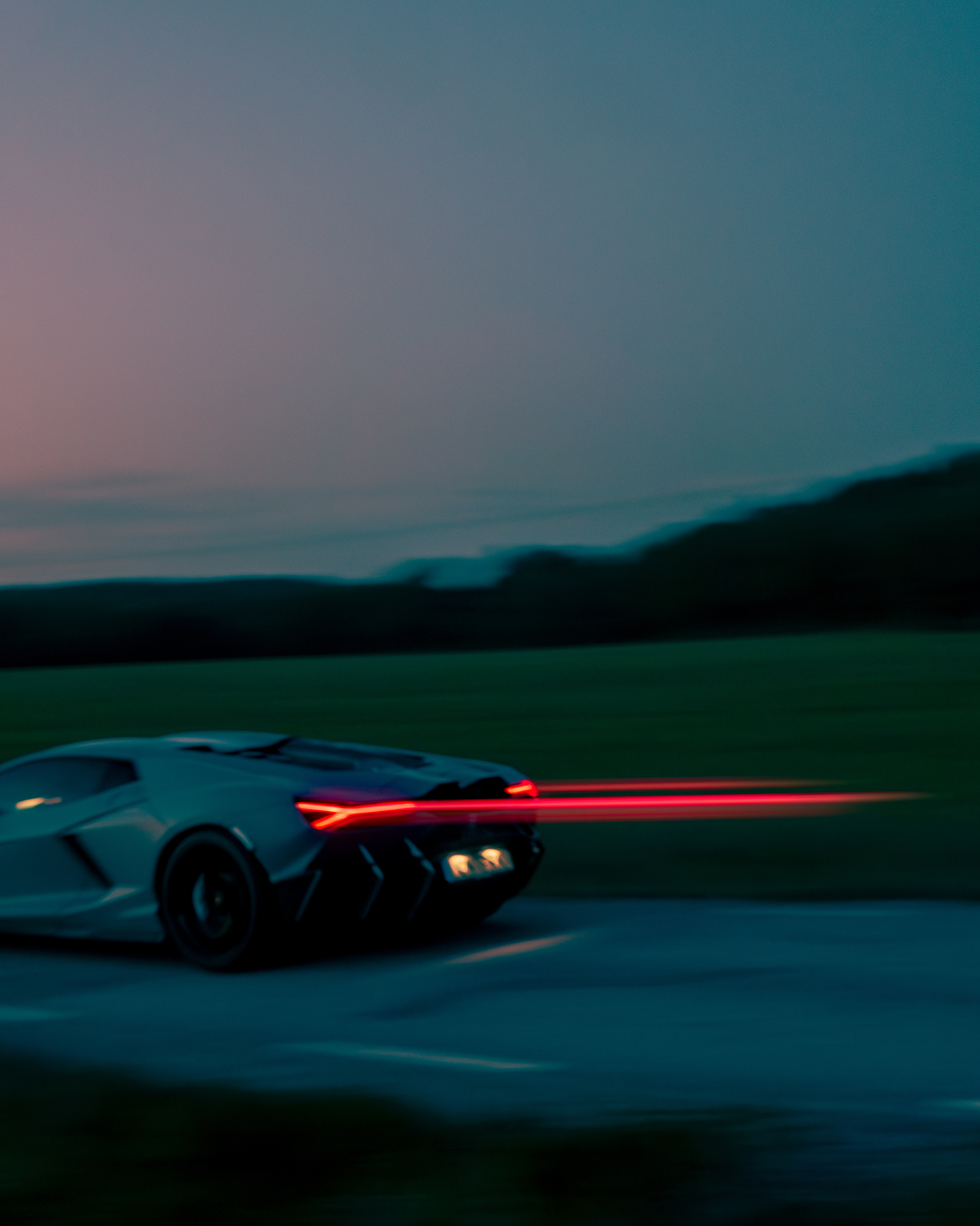 A grey sports car driving on a road at dusk, with a bright red light bar effect extending from the rear taillights against a dark landscape and blue sky.